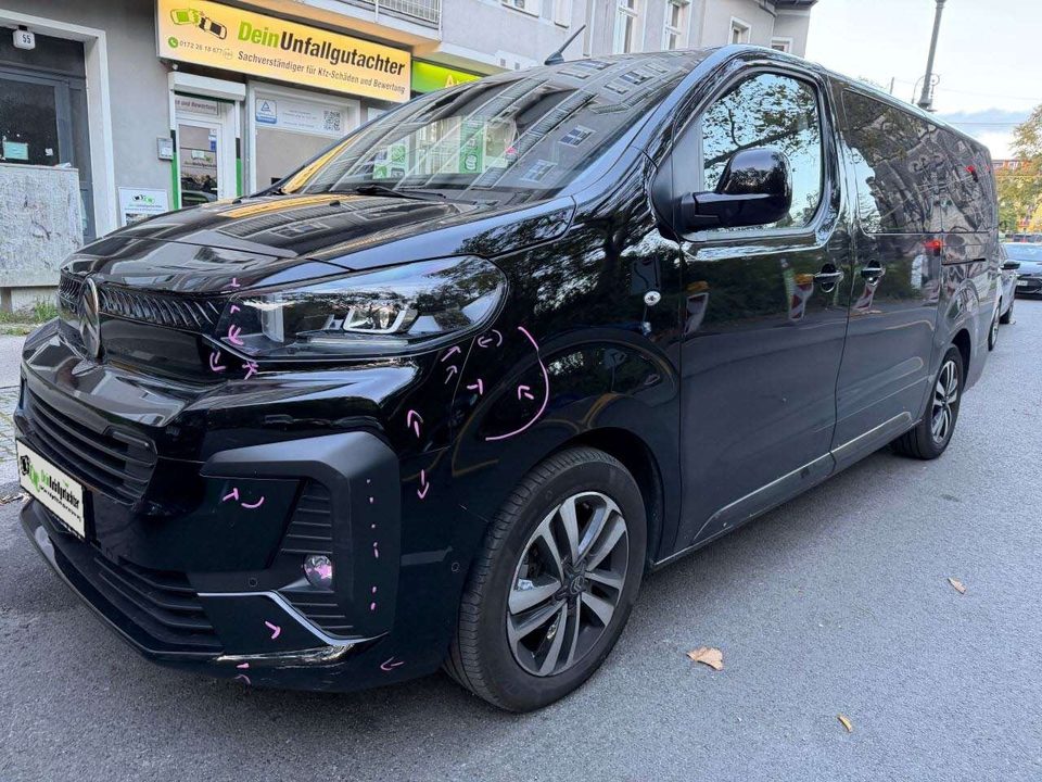Schwarzer Großraum-Van mit Anstoß vorne links vor dem Büro von Dein Unfallgutachter – die Schäden sind mit rosa Kreide markiert, statt des Kennzeichens ist unser Logo zu sehen